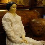 白いレースの洋服を着た陶器製の人形が､壁にもたれかかり遠くを見つめている写真